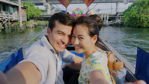 Asian tourist couple having an intimate real selfie moment, taking picture on tour longtail boat, on the chao phraya river water way in Bangkok, Thailand. Slow motion