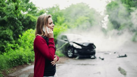 Young woman making a phone call after a car accident, smoke in the background.