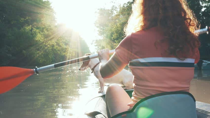 Red-haired young woman paddling on an inflatable kayak with  dog Jack Russell Terrier on river at sunset in beautiful nature, rear view slow motion. Sport. Lens flares in green nature. Travel. Tourism