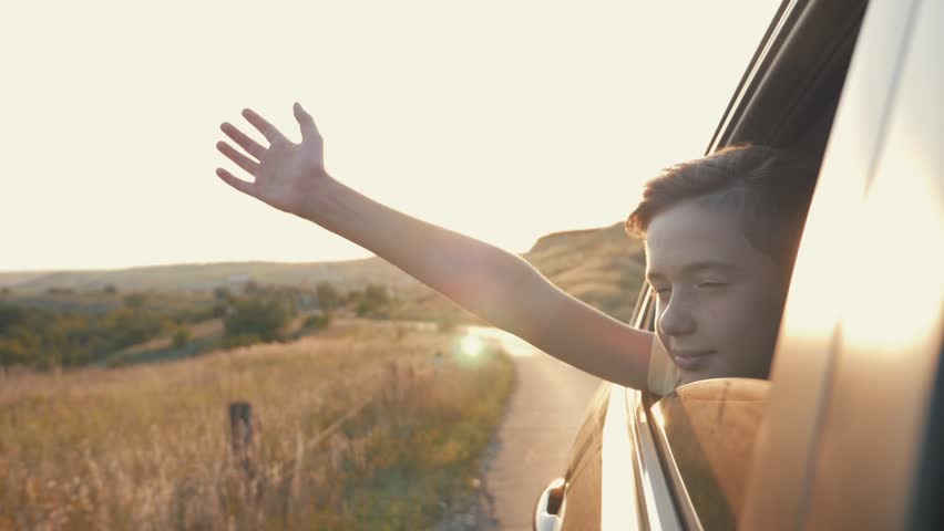 Teen boy looking out the car window and waving his hand. Family travel by car. Hand to wind of freedom. Cheerful boy smiles. Happy family concept. Boy in the car window. The wind of freedom at sunset Royalty-Free Stock Footage #1014453575