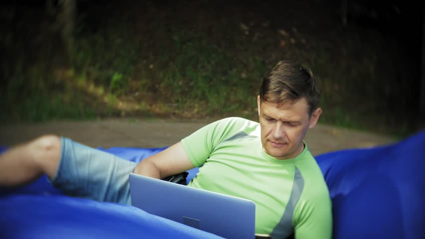 mature man uses a laptop, lying in a lounge area of the park on soft puffs 4k