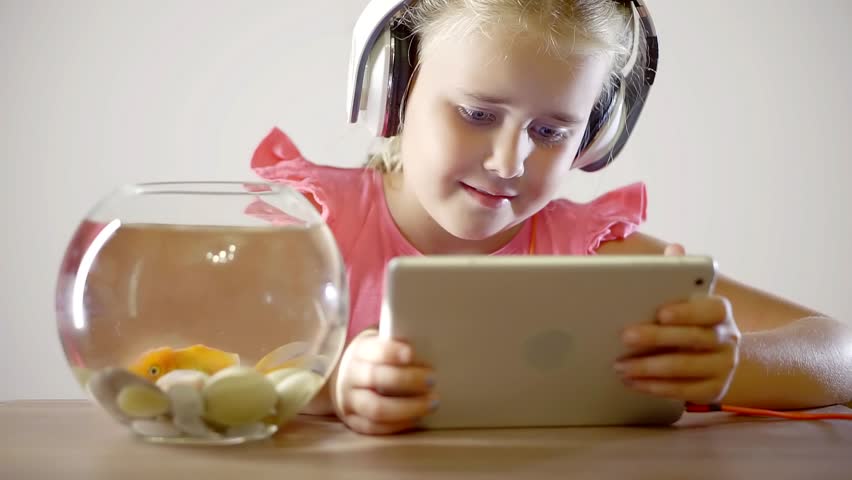 Beautiful girl watching a cartoon on the tablet listening to the sound in the headphones on the table is a goldfish swimming in the aquarium | Shutterstock HD Video #1014463718