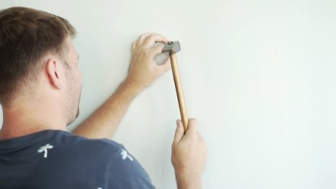 a man is standing by the wall and hammering a nail with a hammer. He knocks on the nail with a hammer, the nail enters the syten.