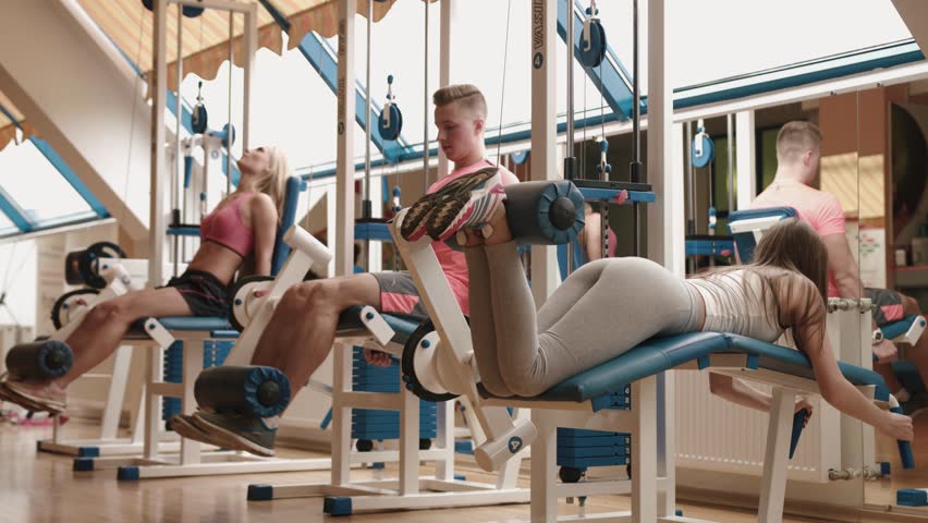 Sport and healthy life style concept. Three young people in sportswears are working out in the gym. They are waming up on the leg curcl trainer. 4k footage.