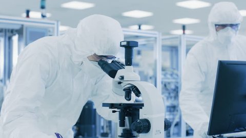 In Laboratory Scientists in Protective Clothes Doing Research, Using Microscope and Entering Data into Personal Computer.