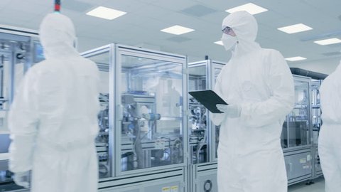 Quality Control Check: Scientist Using Digital Tablet Computer and wearing Protective Suit walks through Manufacturing Laboratory. Product Manufacturing: Pharmaceutics, Semiconductors, Biotechnology.