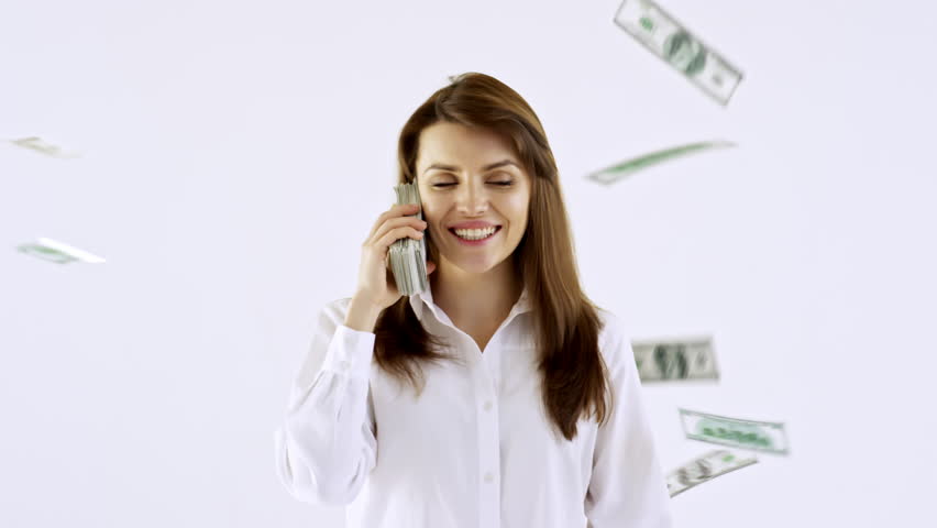 Studio shot of happy young businesswoman standing isolated on white background and using stack of cash as mobile phone as money falling on her from above