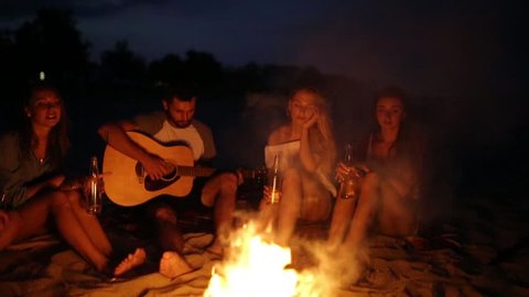 Beach party at sunset with bonfire. Friends sitting around the bonfire, drinking beer and singing to the guitar. Men and women hold glass bottles with beverage singalong, bearded guy playing guitar.