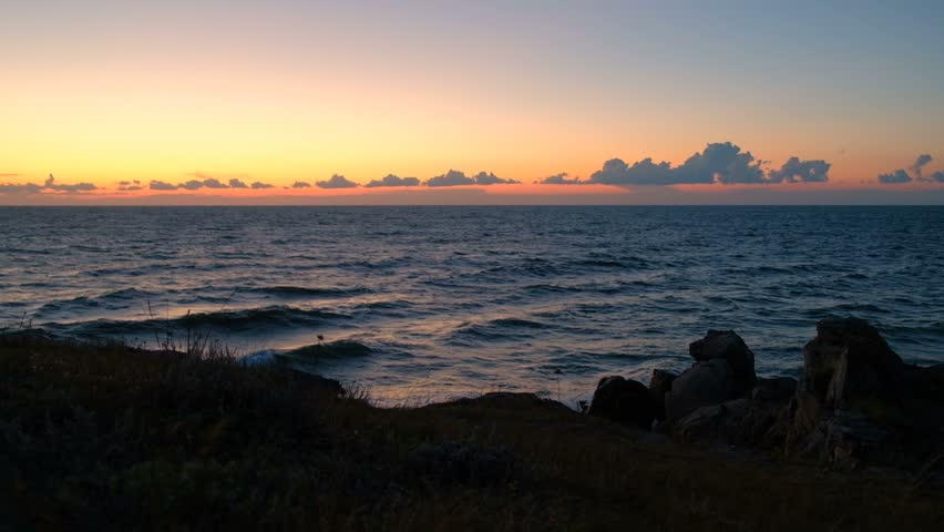 Dramatic coastal nightscape: stunning sunset over water captures twilight atmosphere, painting atmosphere with vibrant orange, pink, and purple radiance. Gentle waves roll and shimmer across offshore expanse, reflecting warm glow from dramatic sky while creating soothing rhythm. Rugged shoreline foreground features texture from sturdy structures and natural elements, framing oceanic composition with depth and contrast. Wispy cloudscape floats delicately during late afternoon, casting subtle shadows that enhance tranquil mood of moment. Soothing illumination characteristic of dusk provides soft glow enveloping entire scene in peaceful ambiance. Horizontal composition showcases expansive aquatic vista with horizon inviting viewers into contemplative infinity. Ideal for travel promotions, lifestyle content, wellness concepts, and environmental campaigns focused on natural harmony.
