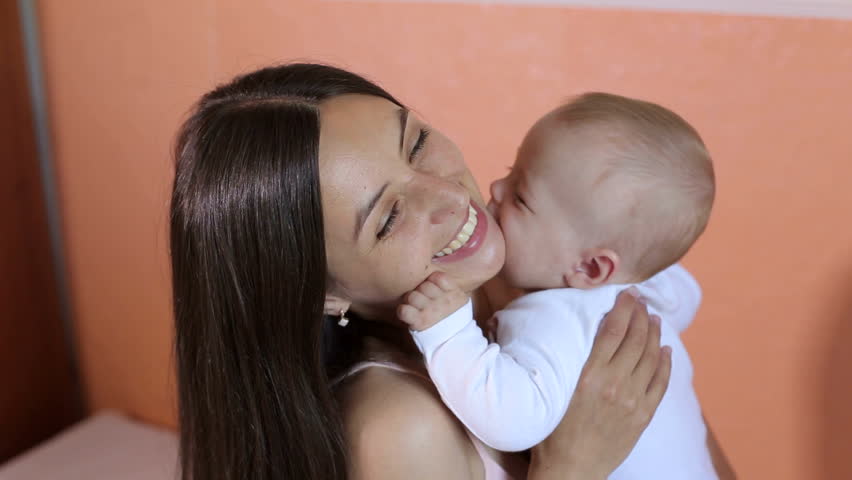 Happy young mom kisses and hugs her newborn baby at home in the bedroom. Family, parenting and child care concept - happy mother kissing adorable baby. | Shutterstock HD Video #1014516917
