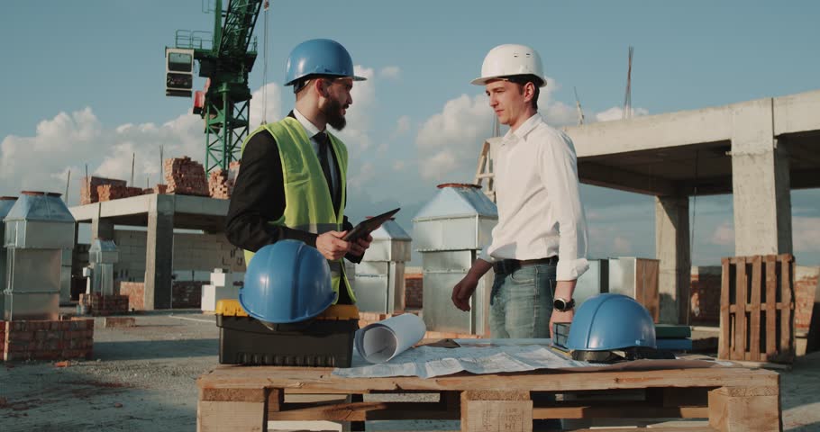 Two young engineers walking through construction site , background big crane , they wearing safety equipment. red epic | Shutterstock HD Video #1014527384