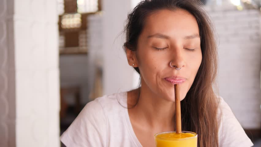 Young Attractive Mixed Race Woman Drinking Fresh Tropical Mango Smoothie Shake Juice Using Zero Waste Eco Friendly Bamboo Straw in Vegan Restaurant. 
