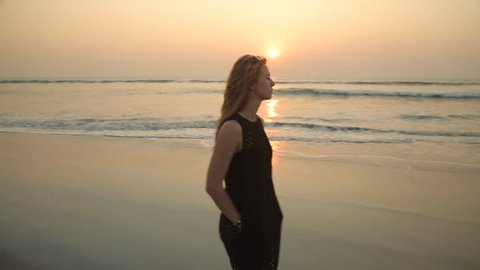 A young attractive blonde girl in a black short dress walks along the beach at sunset in a bad mood sad offended. Stabilized following camera.