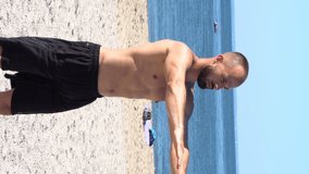 Vertical Video Frame - Young athletic build man exercising on beach waterfront with ocean in background. Free standing squats to strengthen core muscles on sand not wearing shirt