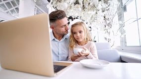 Cheerful father sitting with daughter in cafe while using laptop computer and drinking tea