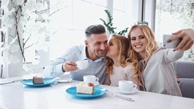 Happy lovely family sitting together by the table in cafe while making selfie on smartphone