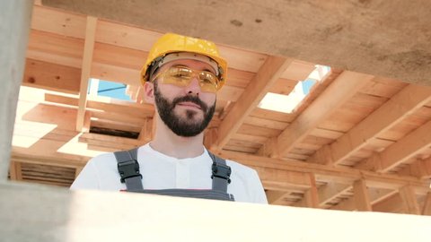 Portrait of a male builder in overalls and a yellow helmet on the roof of a frame house. Screws with a screwdriver.