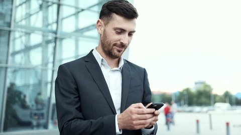 Young Attractive Businessman Standing near Big Modern Office Building. Typing a Message on his Smartphone. Looking Satisfied. Bearded Man Wearing Classical Suit. Business Lifestyle.