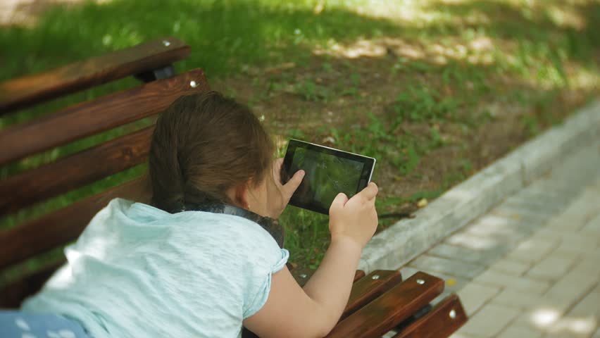 Little fat girl with a tablet PC and headphones sitting on a bench listening to music or watching a video in a summer park 4k