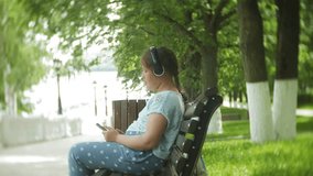Little fat girl with a tablet PC and headphones sitting on a bench listening to music or watching a video in a summer park 4k