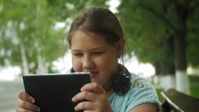 Little fat girl with a tablet PC and headphones sitting on a bench listening to music or watching a video in a summer park 4k
