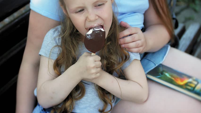 Two little girls are sitting on a wooden bench in a city reading a book and eating ice cream, the background of a city park 4k