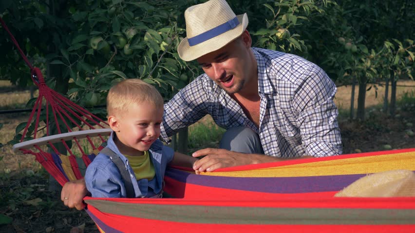 childhood, young father plays with his son in a hammock between rows of trees at the apple garden