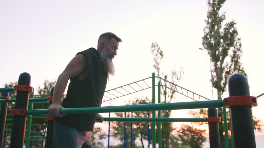Fit handsome middle aged man with long gray beard doing push ups on parallel bars on sports ground with sea background
