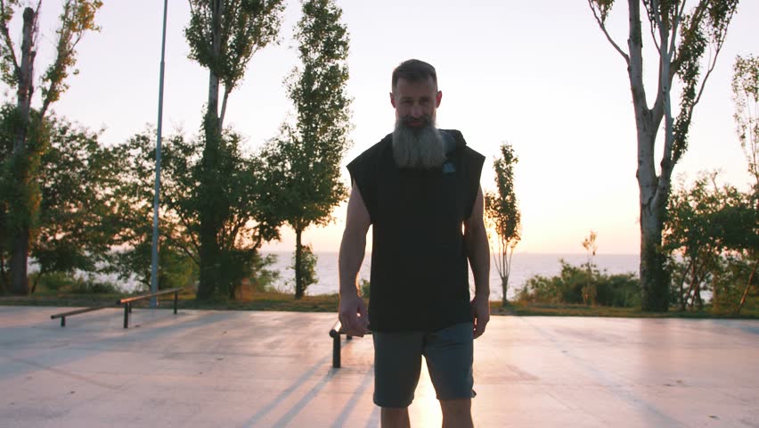 Fit handsome middle aged man with long gray beard doing pull-ups on horizon bar on sports ground with sea background