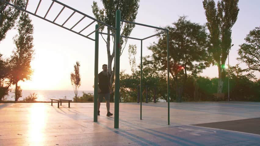 Fit handsome middle aged man with long gray beard doing pull-ups on horizon bar on sports ground with sea background