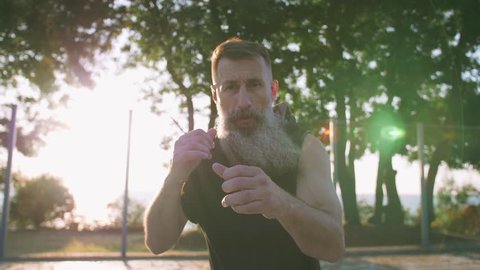 Middle aged man with long gray beard boxing with shadow on basketball court during sunrise, slow motion