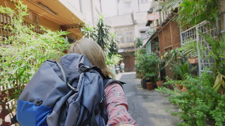 Young couple holding hands blond tourist back packer woman leading boyfriend walking down Asian Bangkok street to explore city. POV travel budget concept. 