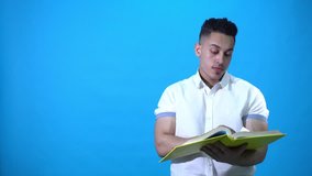 Young man wears a shirt holding a yellow book turning the pages than smiling, on blue Chroma