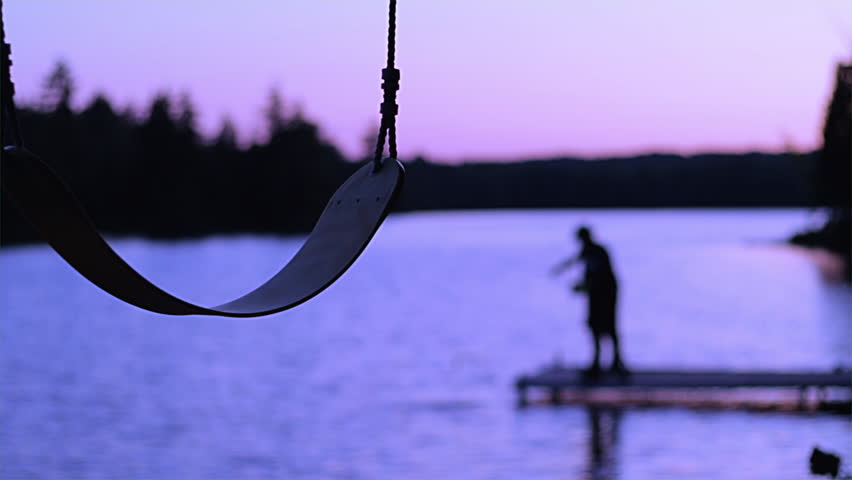 Swing set in silhouette with blurry background of people fishing off a dock on a lake in the background and trees on the horizon. Summer sunset at a cottage. Beautiful sunset in the summer camping. 