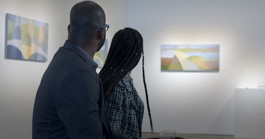 MS African American man and Hispanic woman discuss paintings in gallery during art opening reception. Slider movement with couple in front-sided profile, art soft focus in background