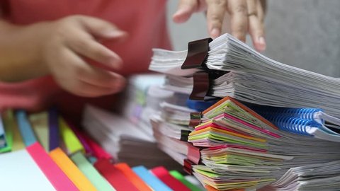 Asian teacher hands is searching for student's homework assignments archive with colorful papers on table to make a check and inspect. Stack of paperwork and reports. Education and business concept.