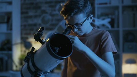 Smart young boy with glasses looking through a telescope in his bedroom and learning astronomy, education and discovery 