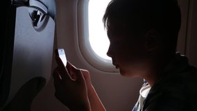 Closeup view of cute profile of young kid sitting in economy class seat in plane near illuminator using smartphone to play video games during flight. Real time full hd video footage.