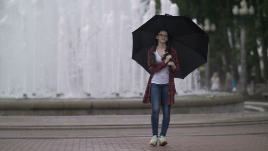 girl with an umbrella walking in the park on a background of a fountain, looking at the camera