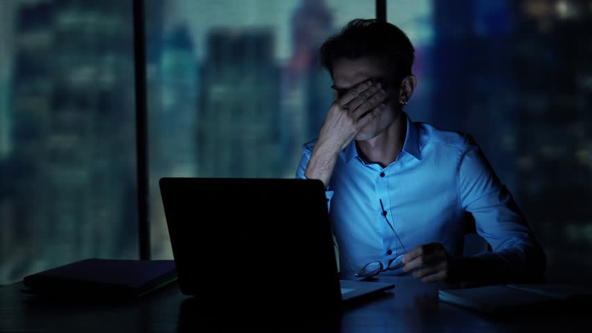 Tired young man working on a laptop late night in the office. Sleepy Businessman sitting at desk in dark office