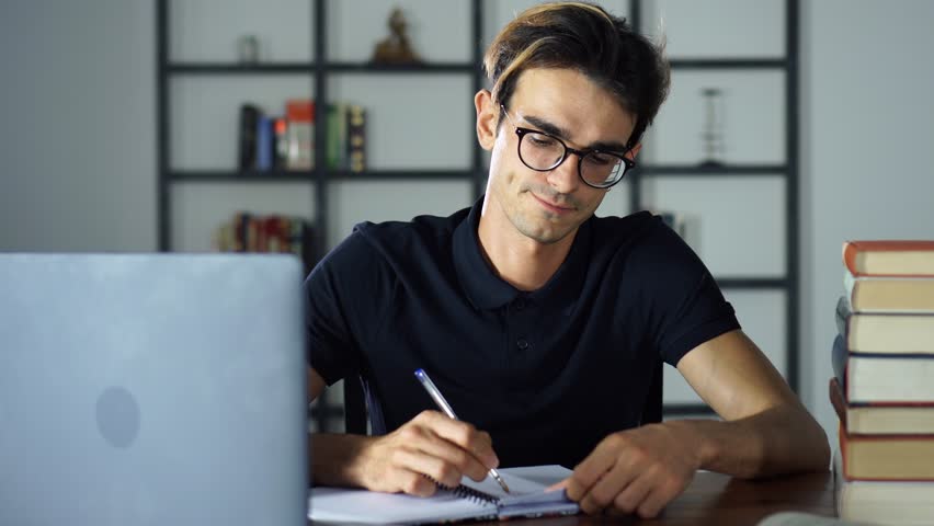 Portrait of Young man sitting at desk, using a laptop, Writing at the notebook, he is studying a book and using a laptop | Shutterstock HD Video #1014621914