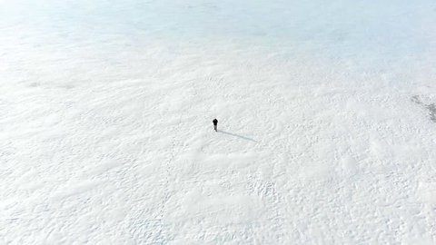 Aerial shot following lone man walking on snowy field in the middle of nowhere. Drone shot of ice tundra with stranded person lost in isolated cold environment. No hope of rescue in snowy disaster.