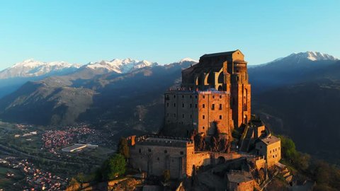 Aerial: drone flying at old medieval abbey perched on mountain top, stunning background of the snowy Alps at sunrise. Sacra di San Michele (italian) - Saint Michel Abbey (english traslation) - Turin, 