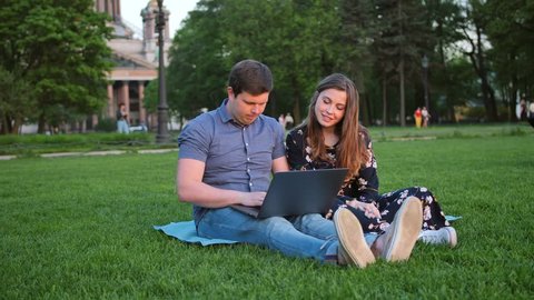 Young Beautiful Couple Looking at Laptop and Laughing, Sitting in Park in Summer with Amazing Sunlight. 4k evening sunset lighting.