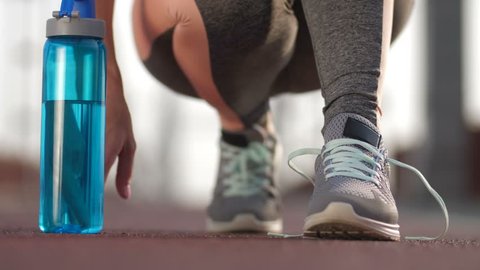 Closeup of female runner tying shoelaces on sneakers, taking sport water bottle and starts running in city. Feet and hands of senior fit lady working out and tying shoes before jogging. Front view