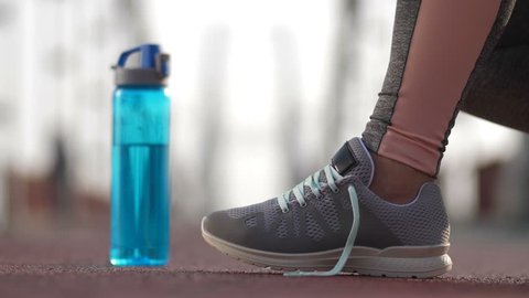 Close-up female runner's feet in sneakers and hands tying shoelaces and taking sport water bottle. Active senior woman running in the morning ties her sports shoes before jogging on bridge. Side view