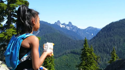 A cute little 12 year old Asian girl pauses in the middle of a mountain hike to enjoy the view and eat a snack on a gorgeous summer day!