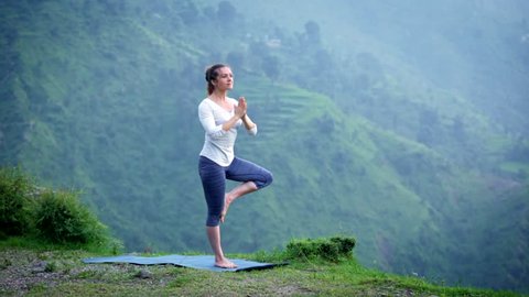 Woman practices balance yoga asana Vrikshasana tree pose in Himalayas mountains outdoors. Himachal Pradesh, India. Panorama