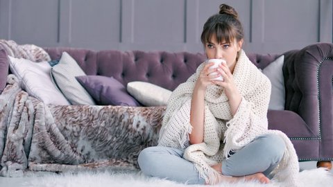 Young woman wrapped in cozy fluffy plaid holding mug sitting on white carpet at home