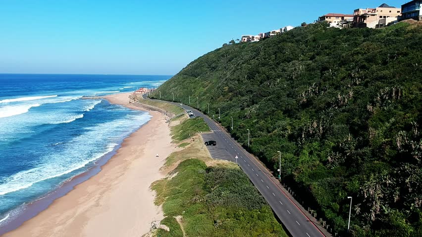 Hovering aerial view of the homes on the beach bluff Durban, South Africa. Sandy beach, blue ocean and crashing waves beside a parking lot along the coast road.